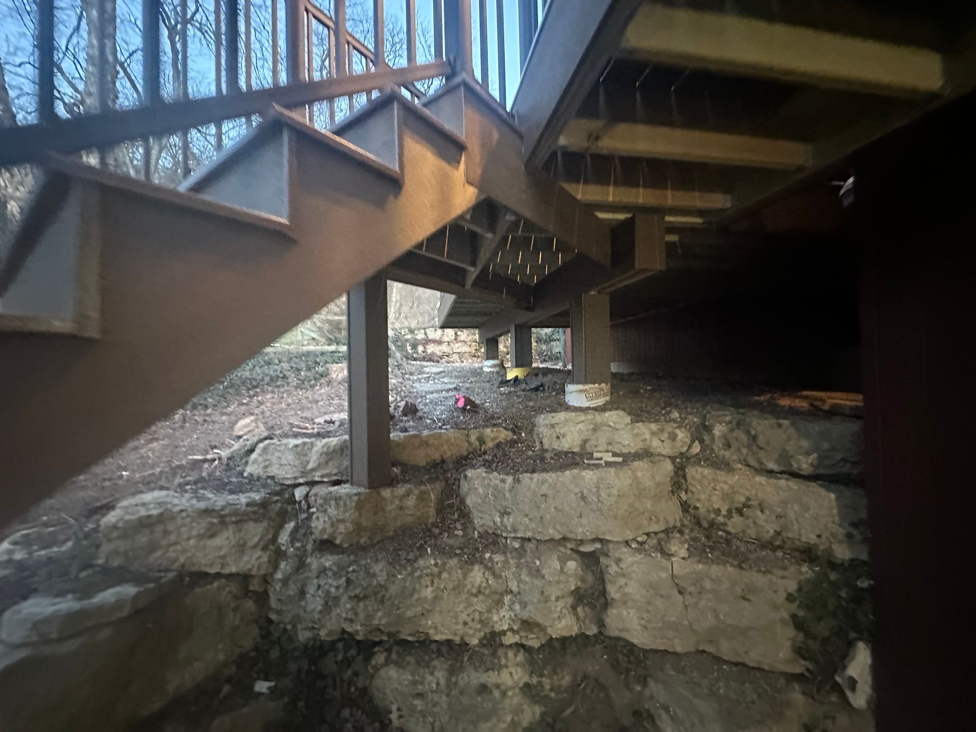 Under a wooden staircase, rough stone ground and concrete support pillars in a dim outdoor space