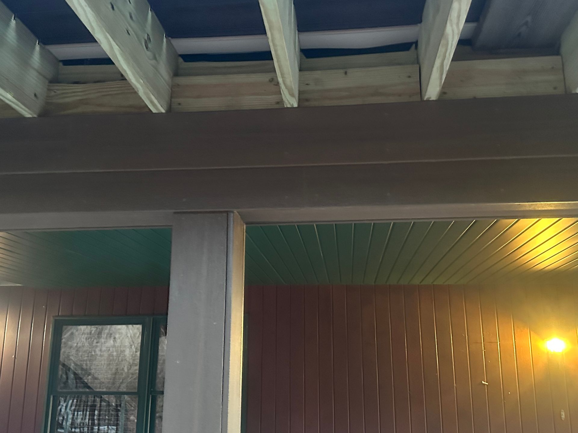 Exterior porch with exposed beams, brown siding, and a lit wall lamp at dusk