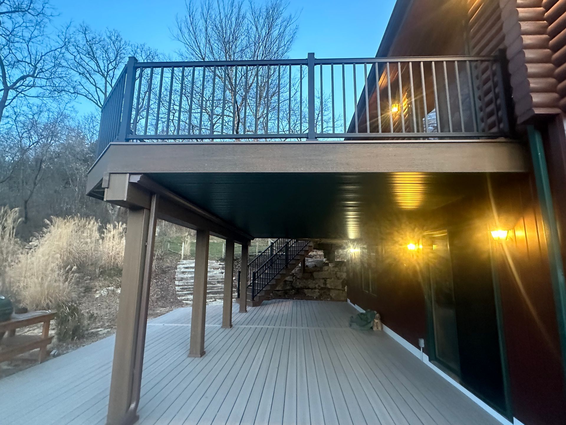 Wooden deck under an upper balcony beside a dark building, lit by warm exterior lights at dusk.