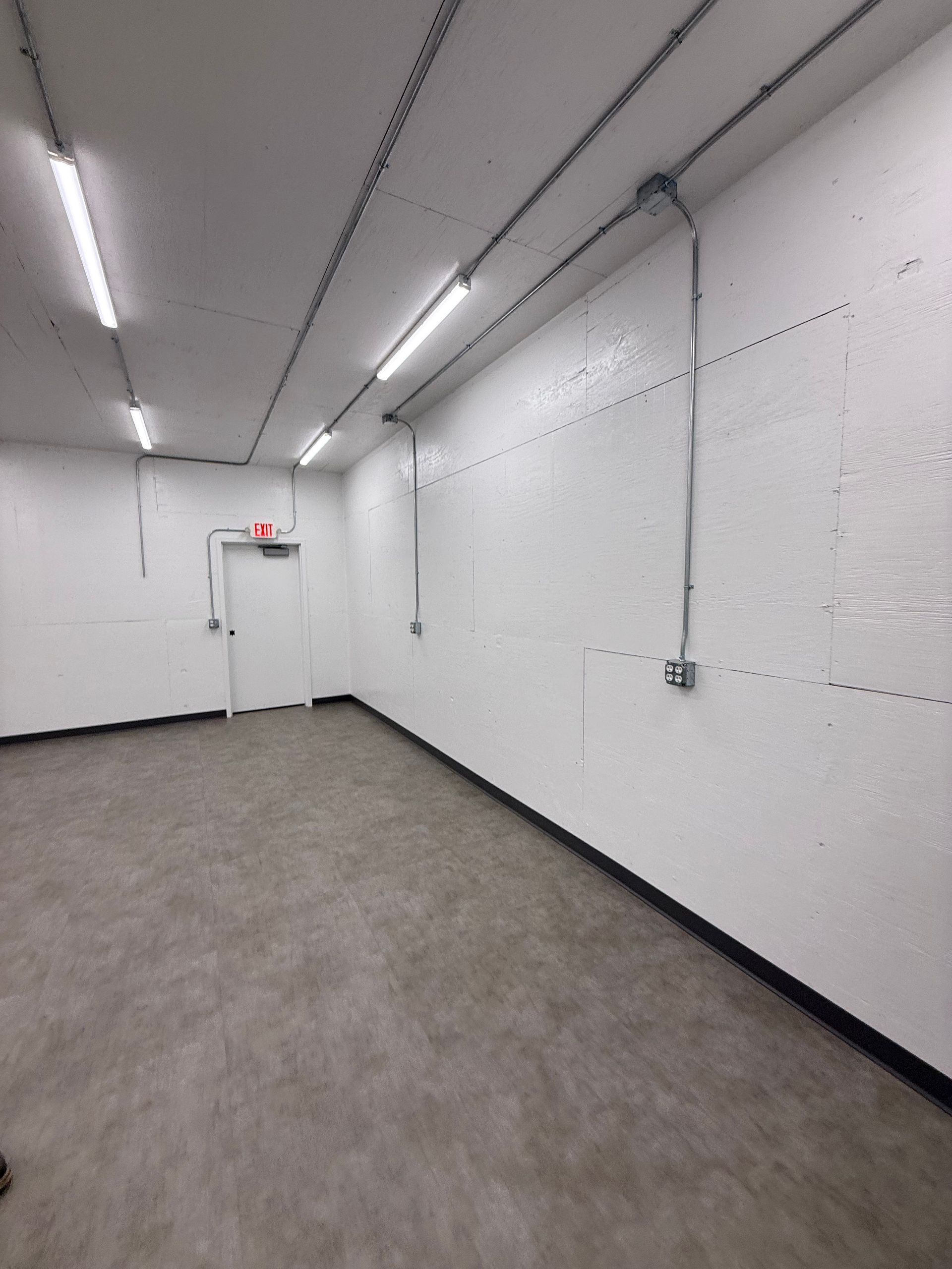 Empty white-walled room with gray carpet and fluorescent ceiling lights, plus a closed door at the far end