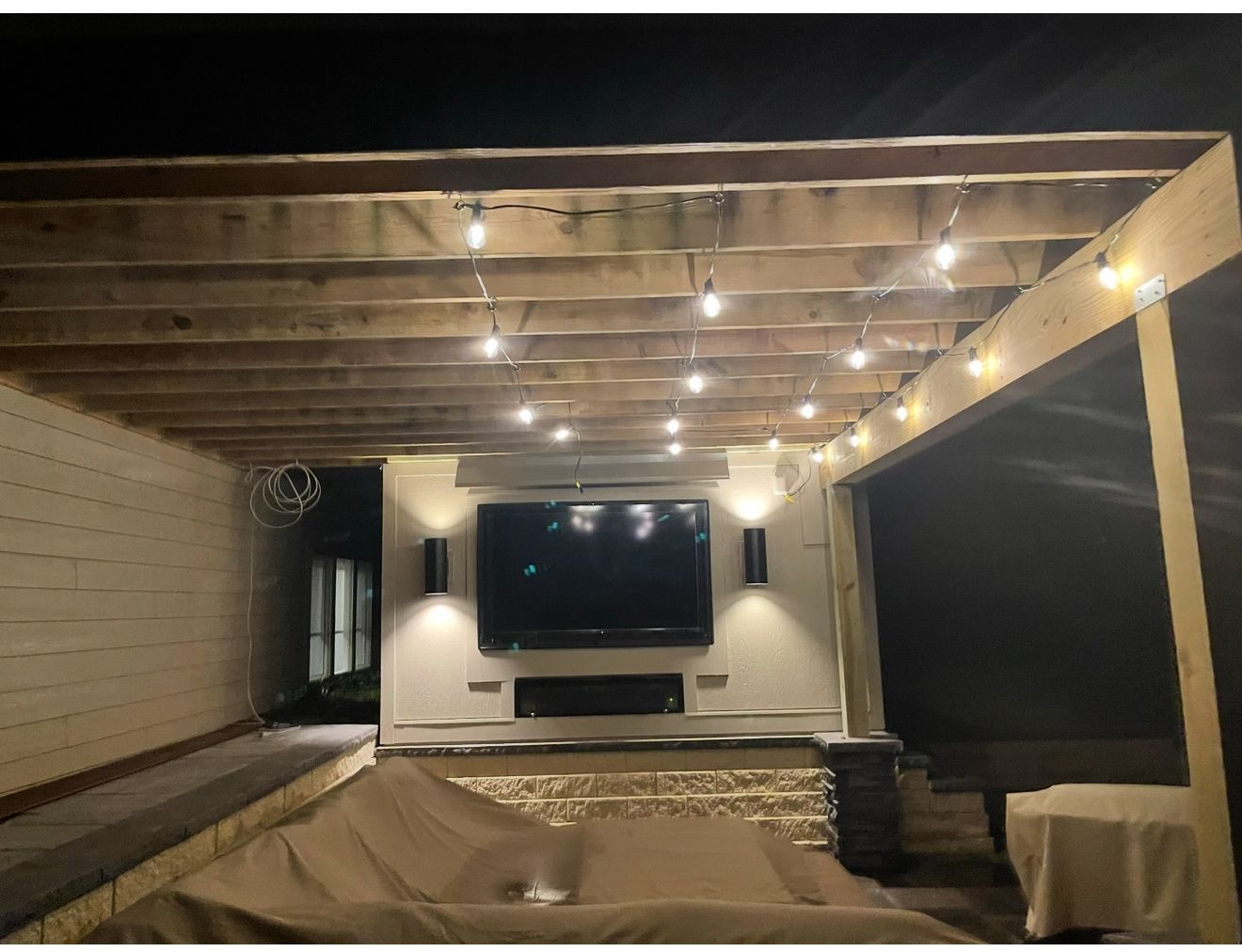 Covered backyard patio with string lights, wall-mounted TV, and a covered hot tub at night