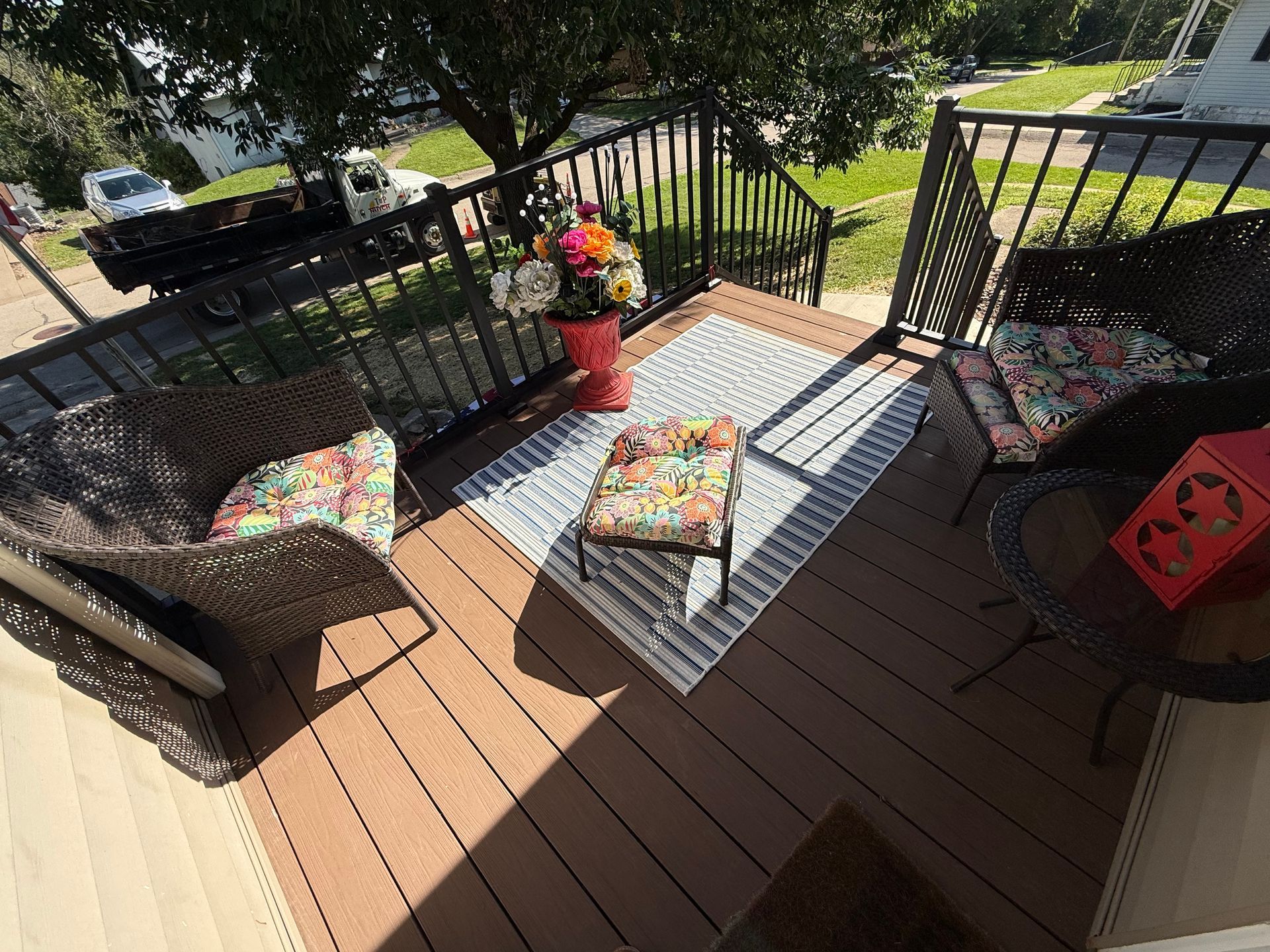 Outdoor deck with two black chairs, patterned cushions, small table, and potted flowers in sunlight