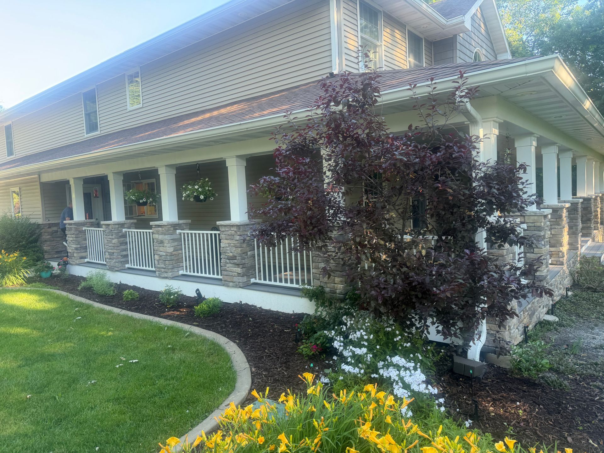 Two-story house with porch, white railing, dark shutters, and landscaped yard with yellow flowers.