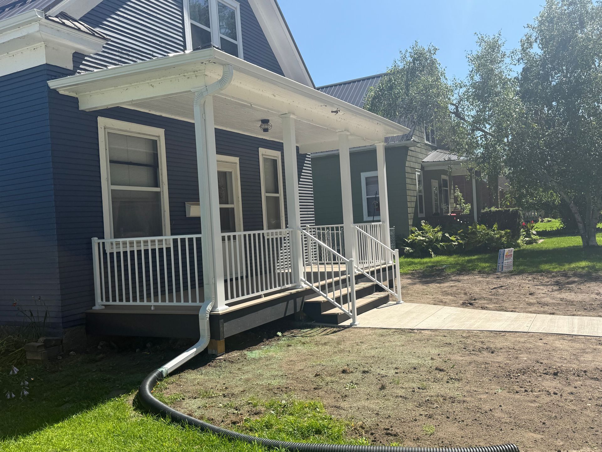 Blue house with white porch railing and steps, beside a dirt yard and trees on a sunny day