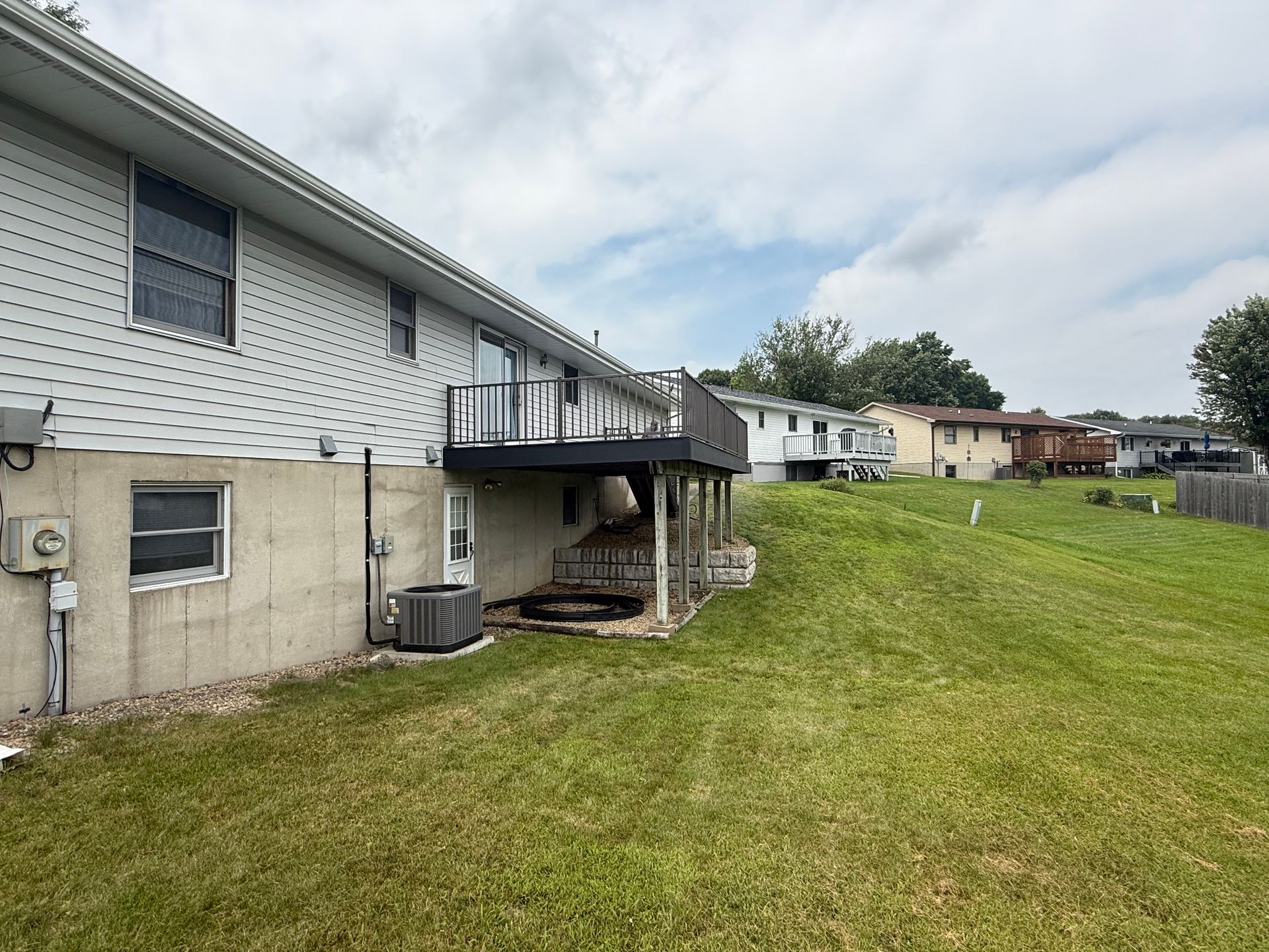 Backyard view of a house with a raised deck, air conditioner, and grassy yard under cloudy sky