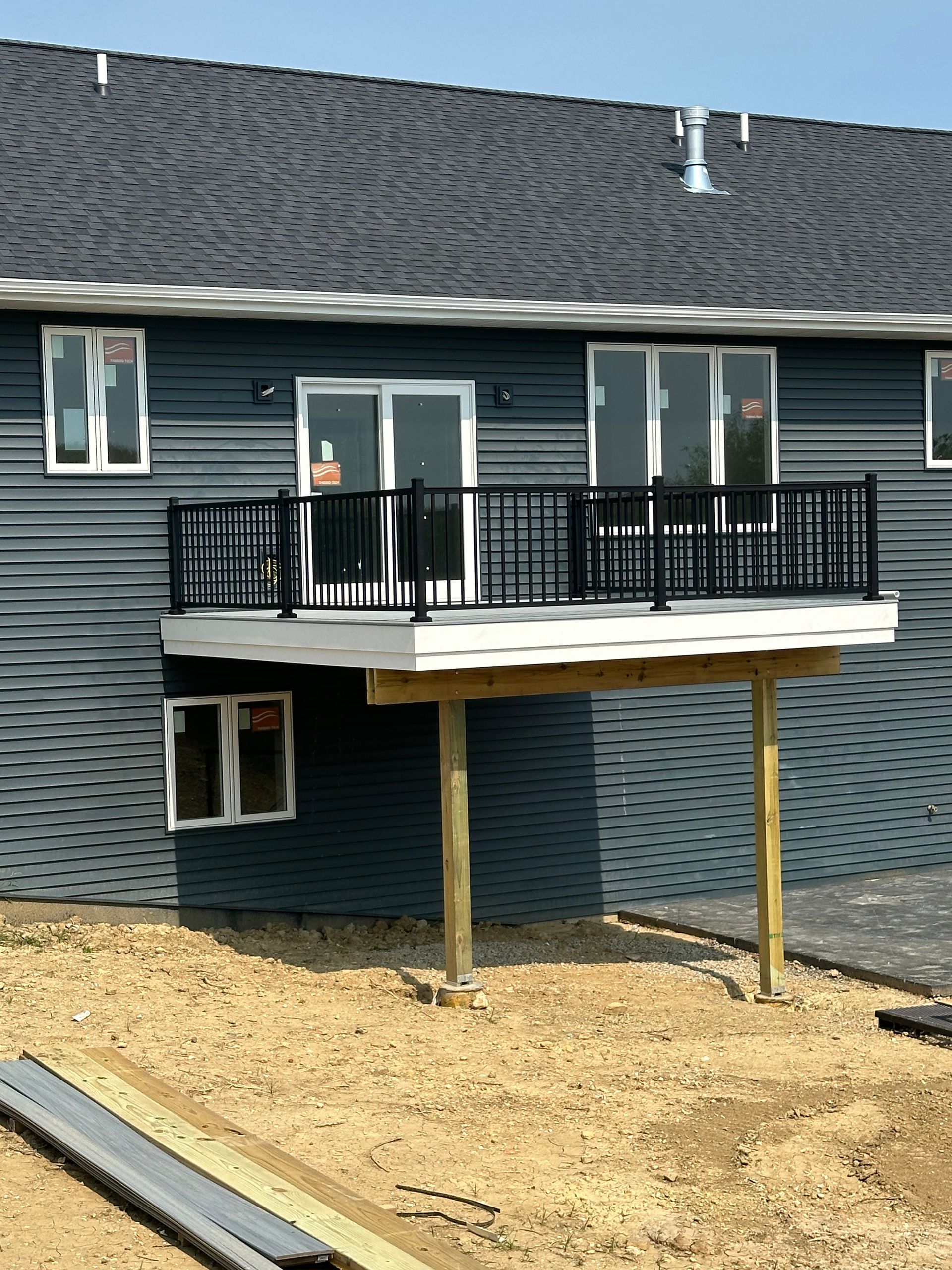 Rear deck balcony with black railing on a blue-gray house, seen over a dirt yard.