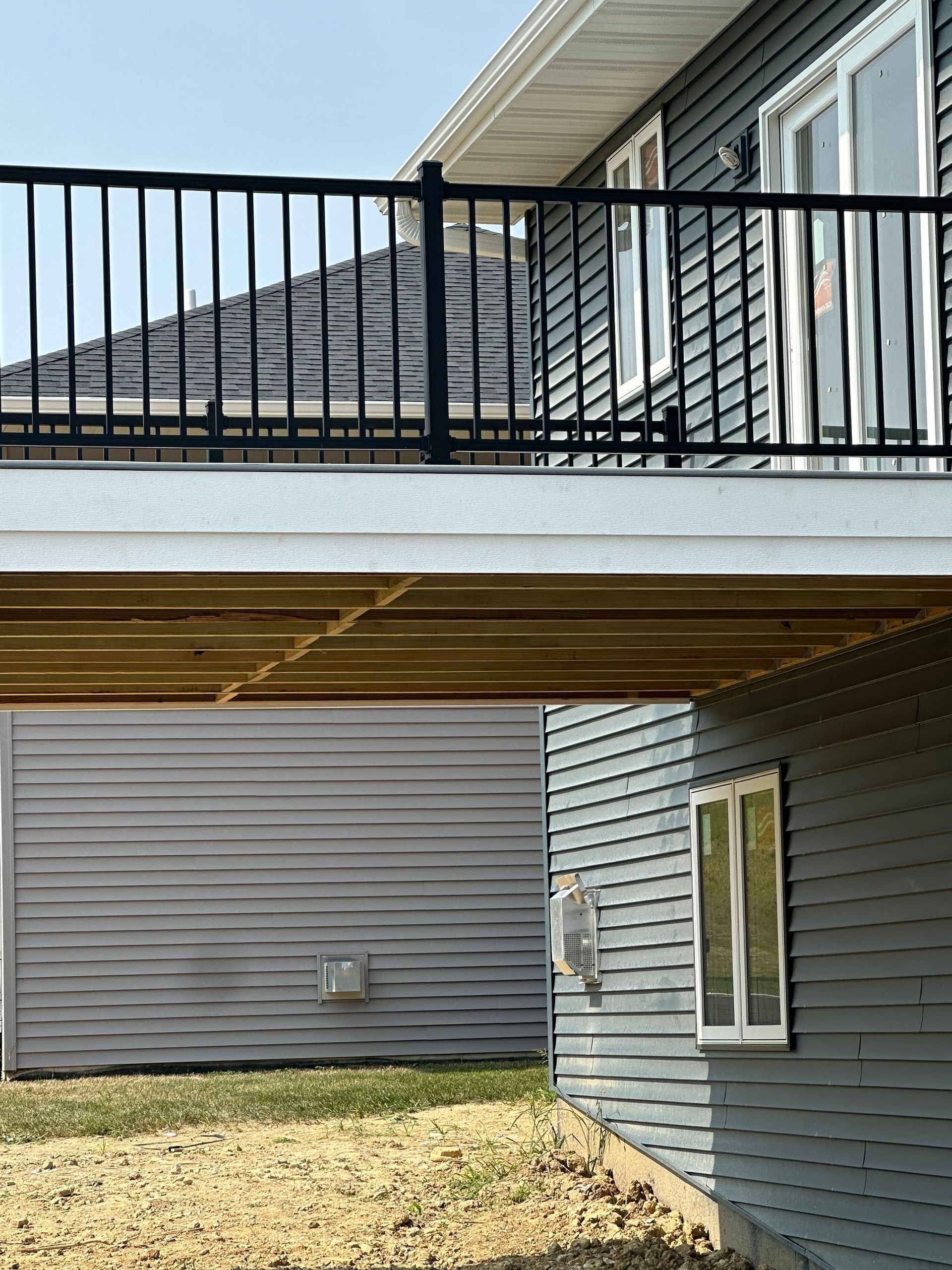 Covered balcony over patio beside gray-sided house, with black railing and windows.