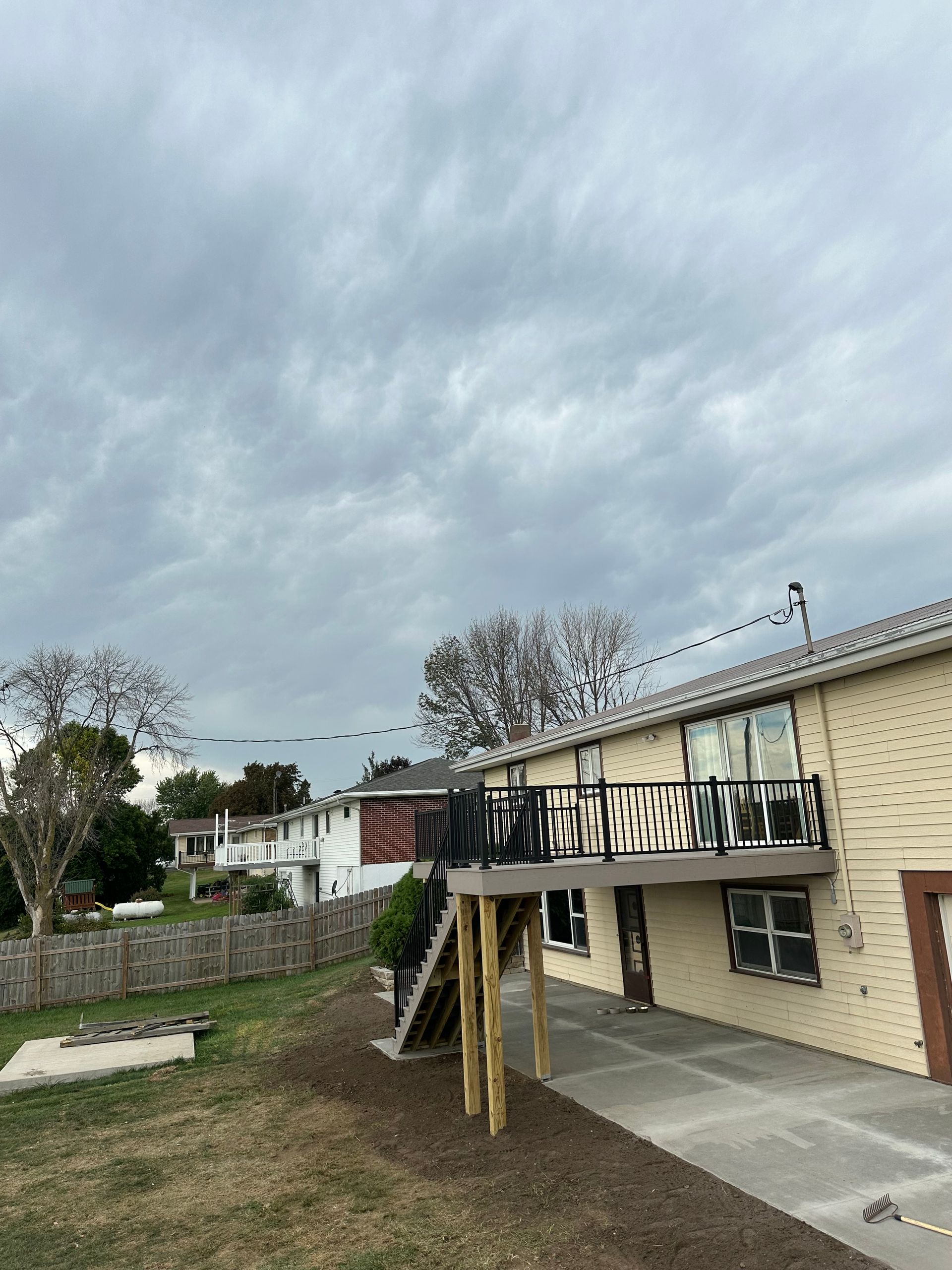 Backyard deck attached to a beige house under a cloudy sky, with a lawn and concrete patio below.