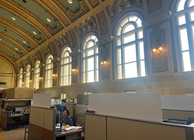 A woman is sitting at a desk in a large room with lots of windows.