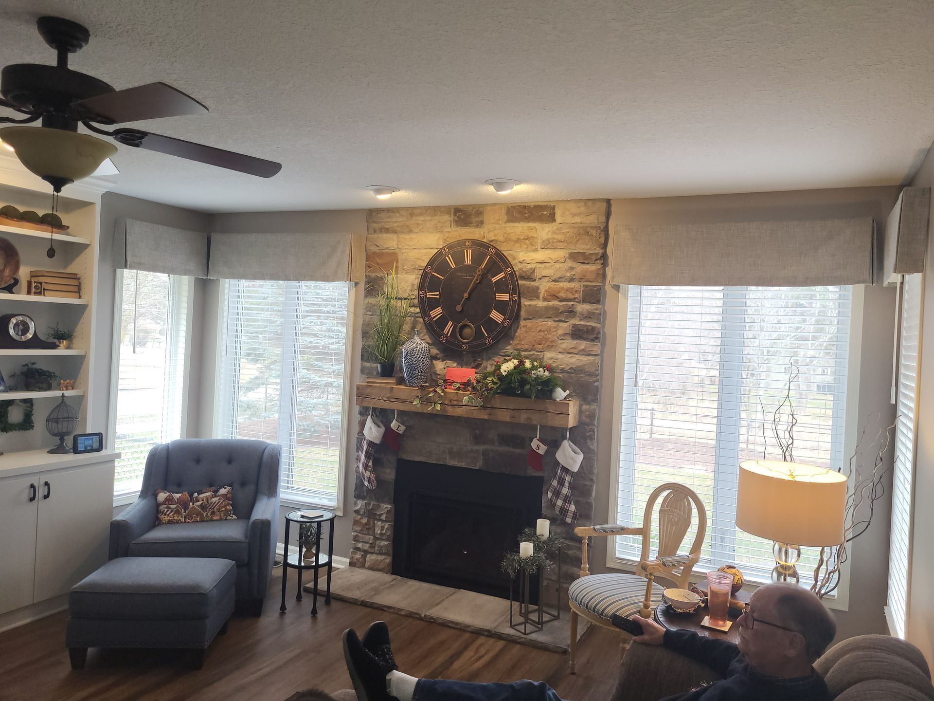 A man is sitting in a living room with a fireplace and a ceiling fan