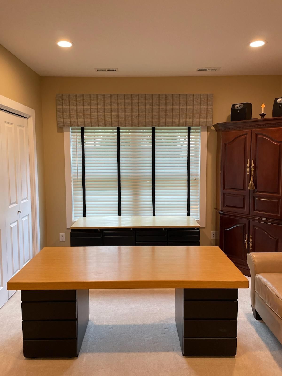 Home office with wooden desk, window blinds, white door, and tall dark cabinet