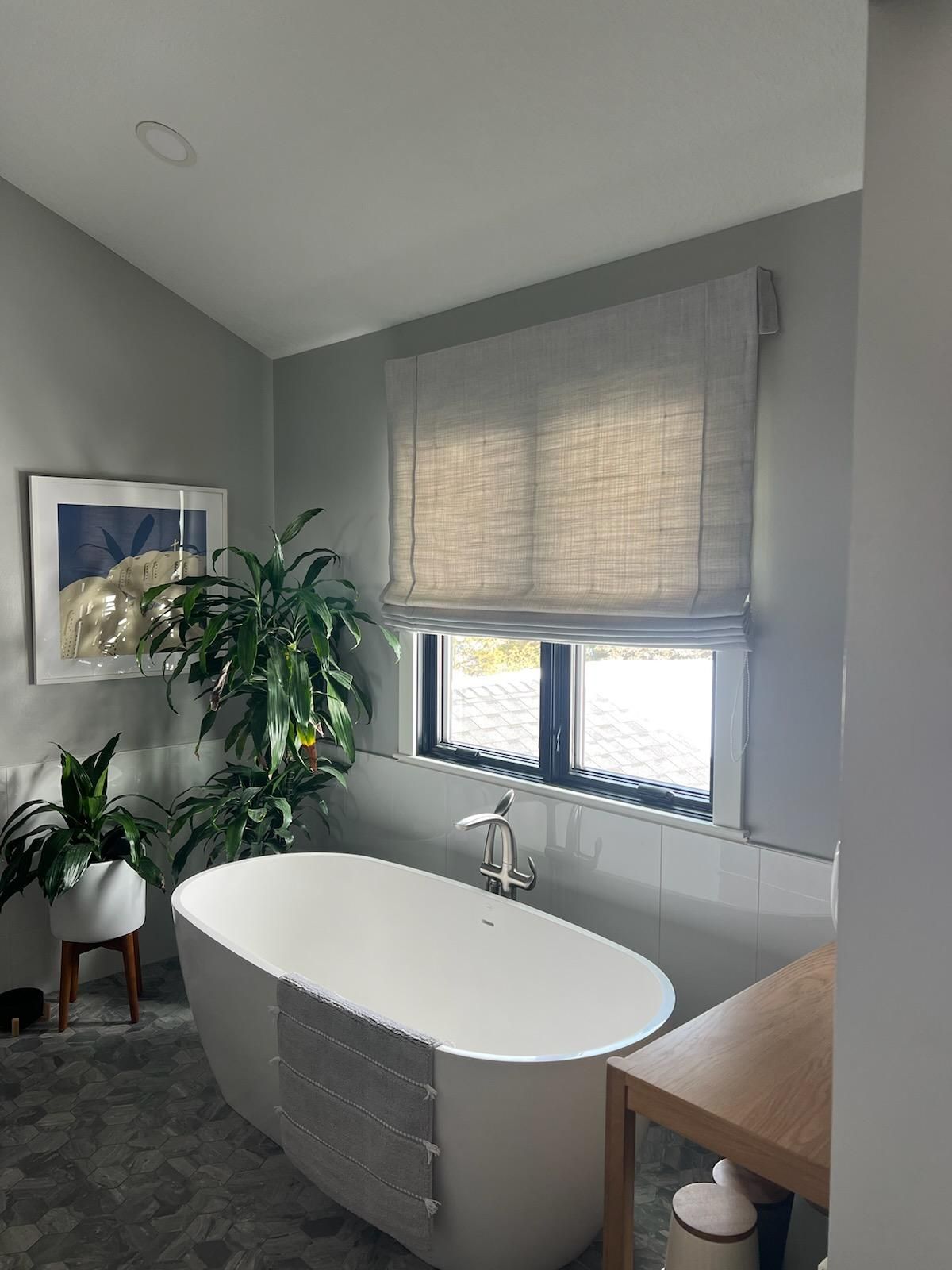 Modern gray bathroom with a freestanding oval tub, window blinds, plants, and a wooden vanity countertop