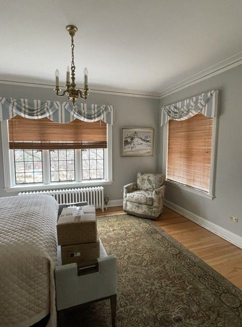 Bright living room with gray walls, bay window blinds, chandelier, rug, and an armchair by the corner window