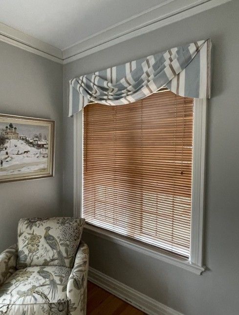 Gray room corner with a floral armchair, framed art, and a window with wood blinds and striped valance