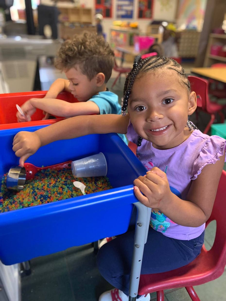 Two children playing with sensory bins at a table, one smiling at the camera.