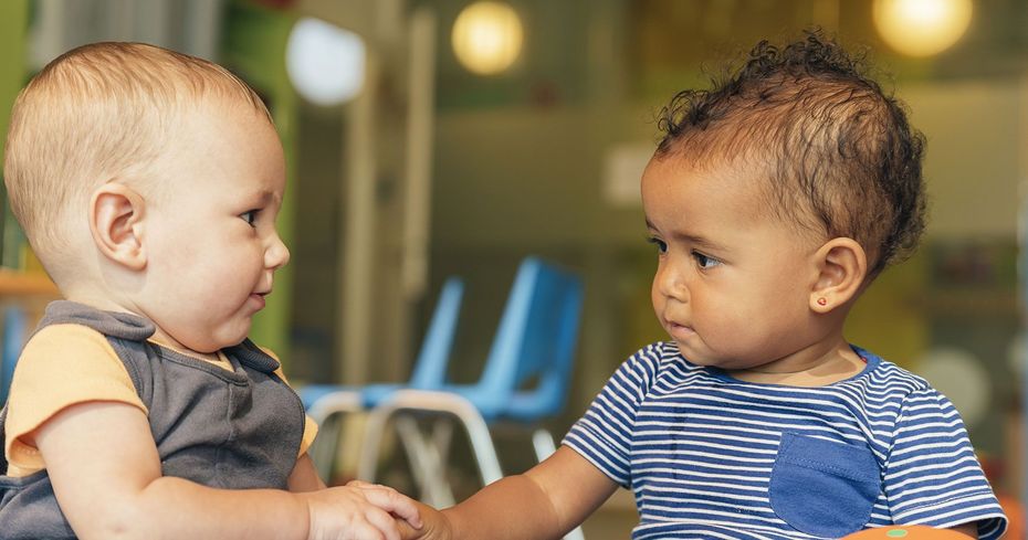 Two babies sit facing each other indoors, holding hands and looking at one another.