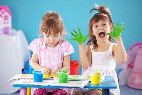 Two children finger-painting at a table, one focused and one smiling with green paint on her hands.