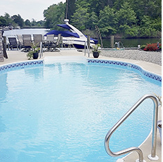 Pool with blue water and tiled edge, dock with boat in background. Trees and patio furniture nearby.