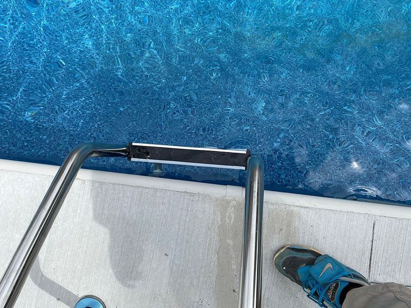 Pool ladder partially submerged in blue water, a foot in a blue shoe near the edge.
