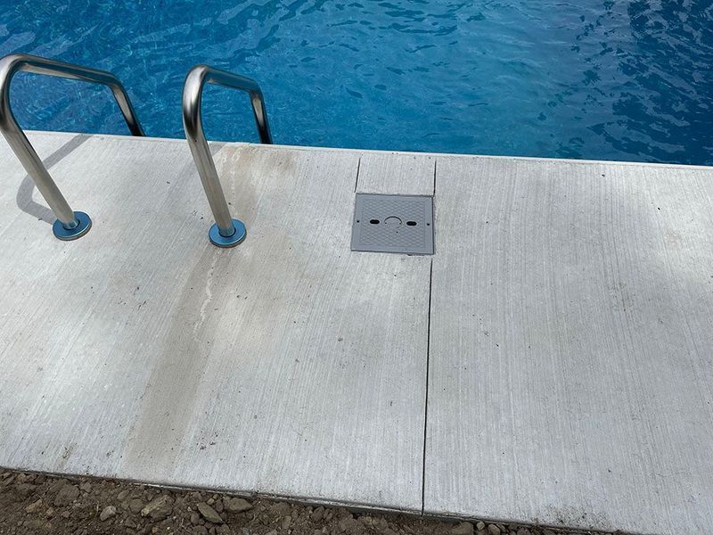 Poolside view with stainless steel ladder, blue water, and gray concrete deck with drain.