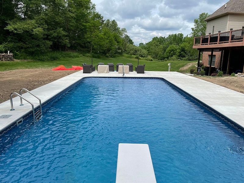 Rectangular in-ground pool with diving board. Concrete deck and chairs. Trees and house in the background.