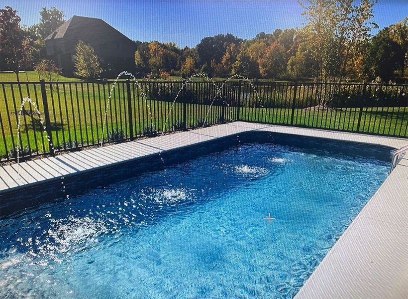 Rectangular blue pool with water fountains, surrounded by a gray patio and black fence, with a grassy area and trees.