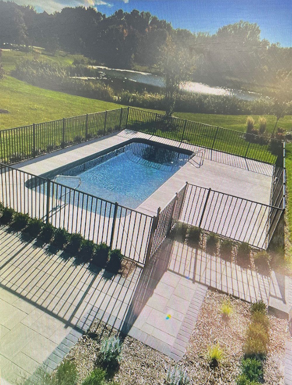 Rectangular outdoor swimming pool surrounded by a black fence and landscaping, set in a grassy yard.
