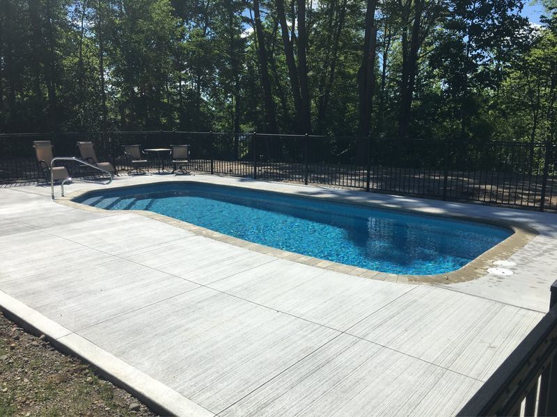 A rectangular in-ground pool surrounded by a concrete patio and a black metal fence, backed by trees.