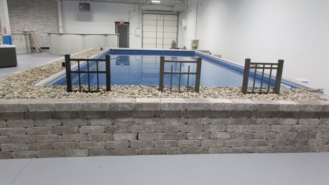 A rectangular pool with black fencing sits within a stone-block raised bed, set in a spacious, white-walled indoor area.