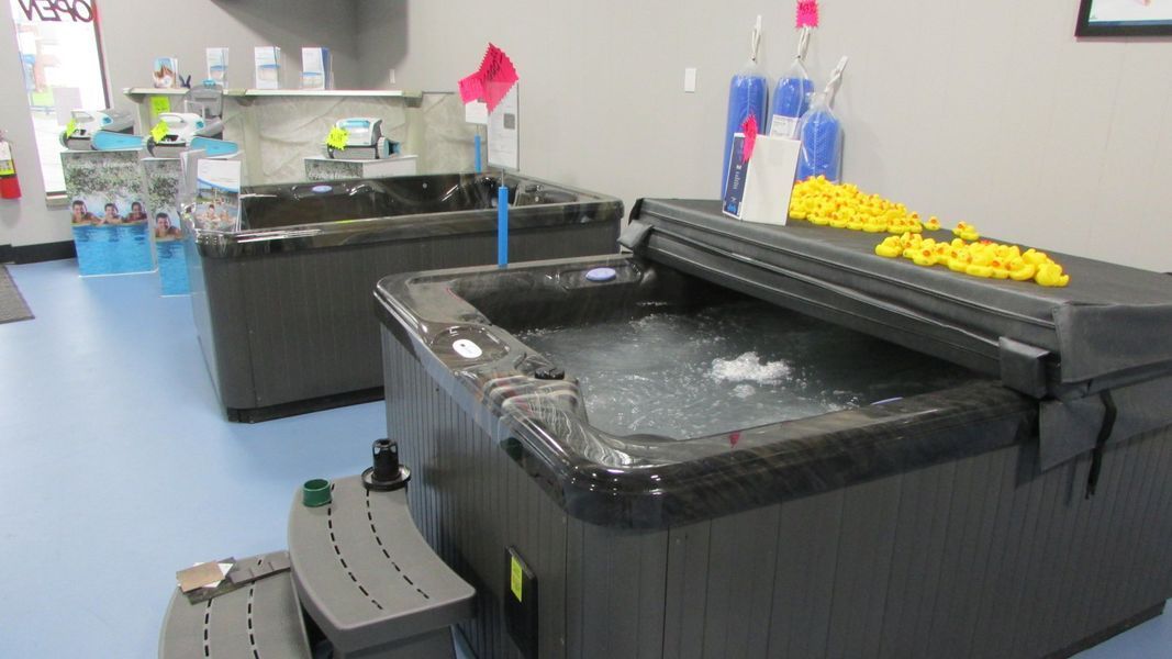 Two dark hot tubs in a showroom, one with the cover partially open and bubbles rising. Blue floor.