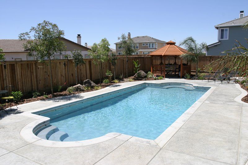 Rectangular pool with steps, concrete patio, wooden fence, gazebo, houses, and blue sky.
