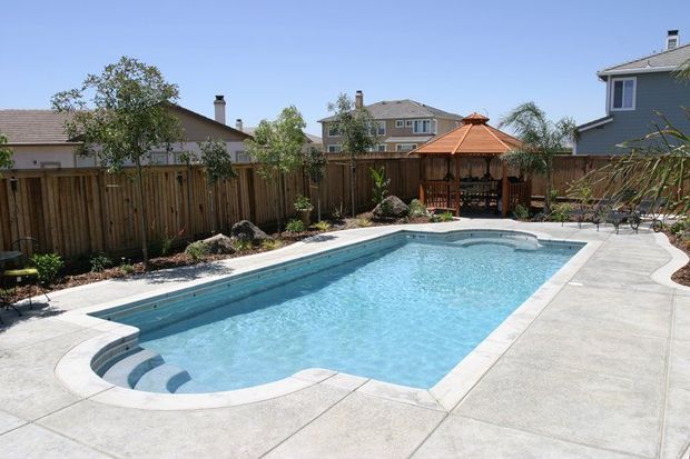 Rectangular pool with steps, concrete patio, wooden fence, gazebo, houses in background.