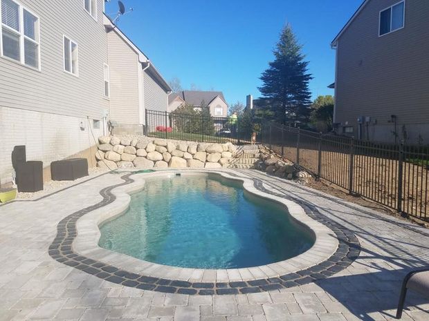 Backyard pool with stone coping and pavers, next to a house with a rock retaining wall and black metal fence.