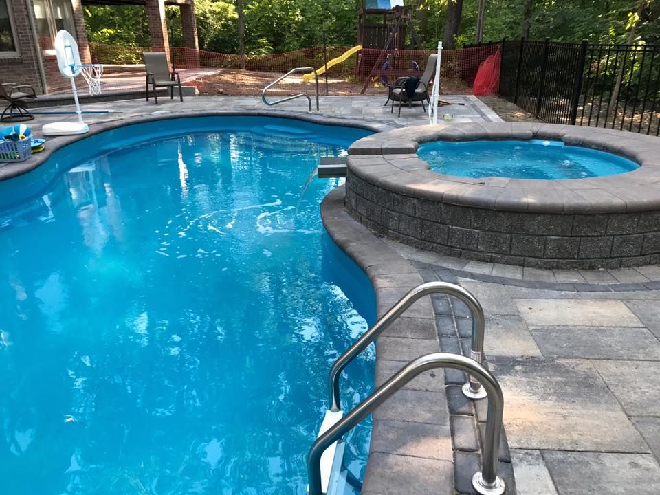 Pool with attached hot tub, blue water, gray stone patio, and stainless steel ladder.