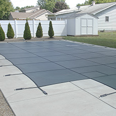 A rectangular pool covered with a gray mesh safety cover, surrounded by concrete and a lawn.