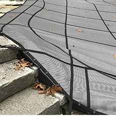A black mesh pool safety cover with straps over stone steps.