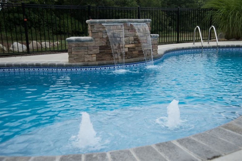 Swimming pool with stone waterfall feature, water fountains, and black fence. Blue water.