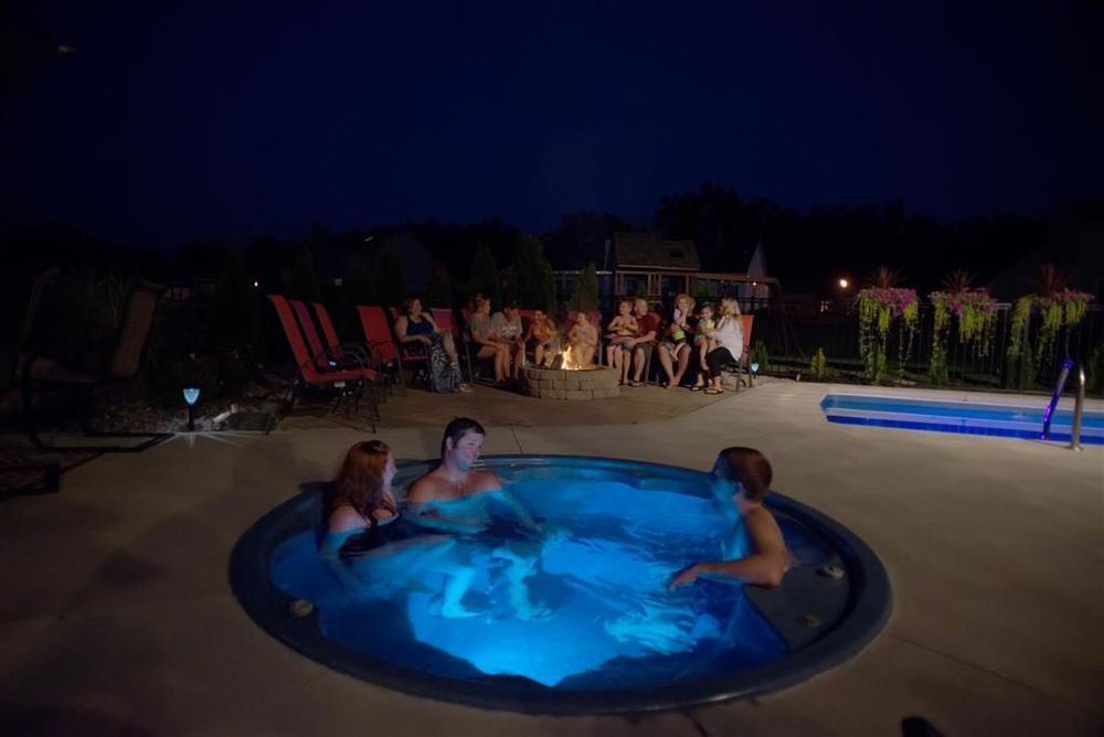 People in a hot tub at night, with a group around a fire pit and a swimming pool nearby.