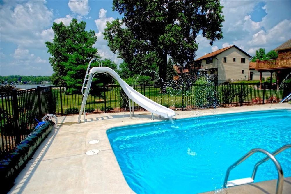 A swimming pool with a white slide, trees, and a house under a cloudy sky.