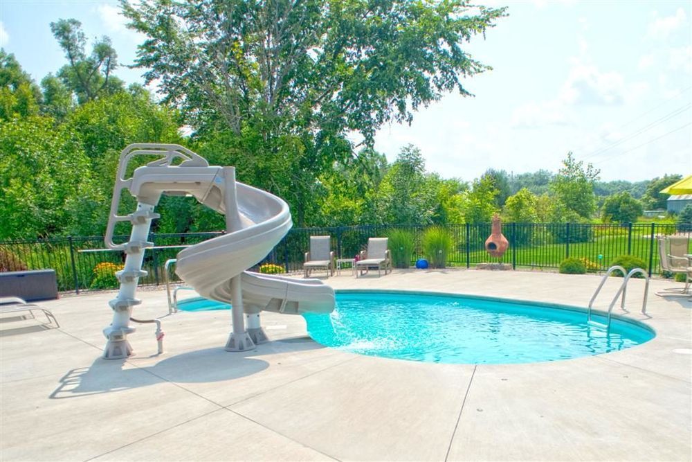 Swimming pool with slide on a concrete patio, surrounded by trees and lawn.