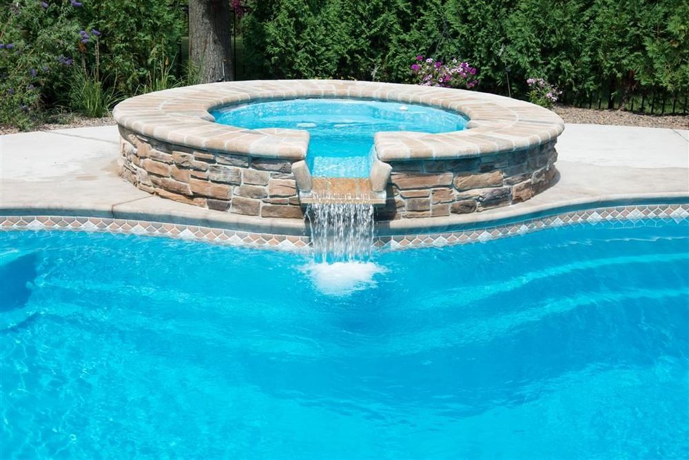 Pool with a circular jacuzzi featuring a waterfall; bright blue water, stone accents, sunny outdoors.