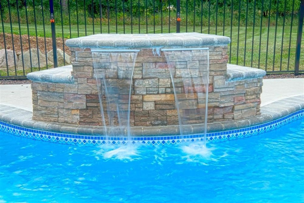 Pool with a stone waterfall feature; water cascading into the blue water, against a black fence.