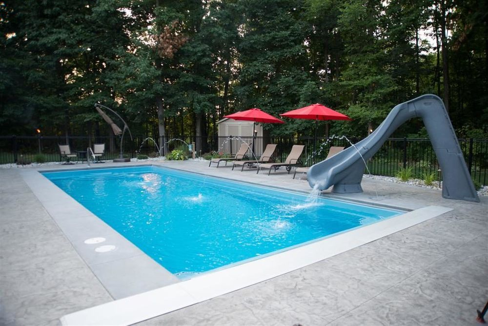 Rectangular pool with slide, lounge chairs, red umbrellas, and water fountains in a wooded backyard.
