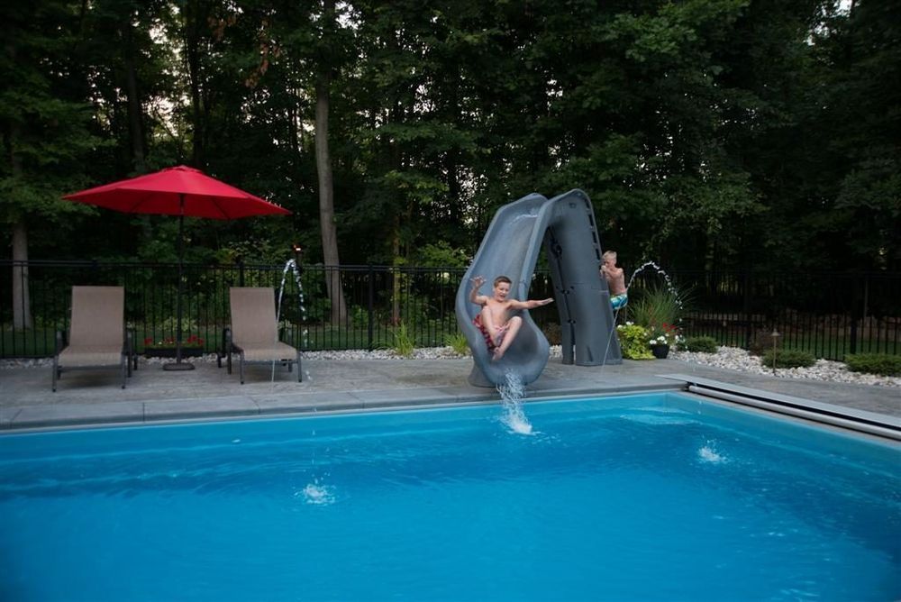Two children slide into a blue pool. Red umbrella, lounge chairs, and trees in background.