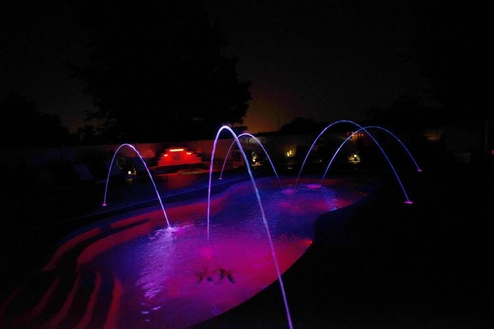 Nighttime view of a pool with glowing, arched water fountains in shades of purple and blue.