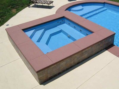 A square spa with brown trim next to a rectangular pool, both blue, on a light tan patio.