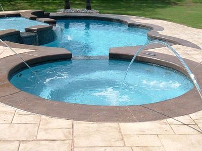 A multi-level pool with clear blue water and stone patio. Water jets spray into the circular hot tub.