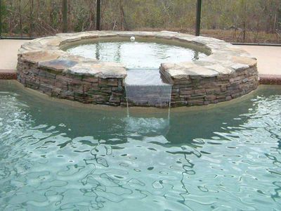 Stone-lined hot tub spilling water into a pool. The hot tub is elevated and circular. Clear water.