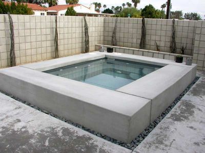 Concrete square spa with glass water, in a backyard setting with a block wall.
