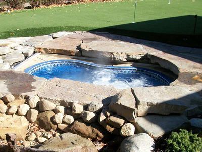 Hot tub surrounded by rocks, set in a backyard with a putting green. Blue tiles inside, water bubbling.